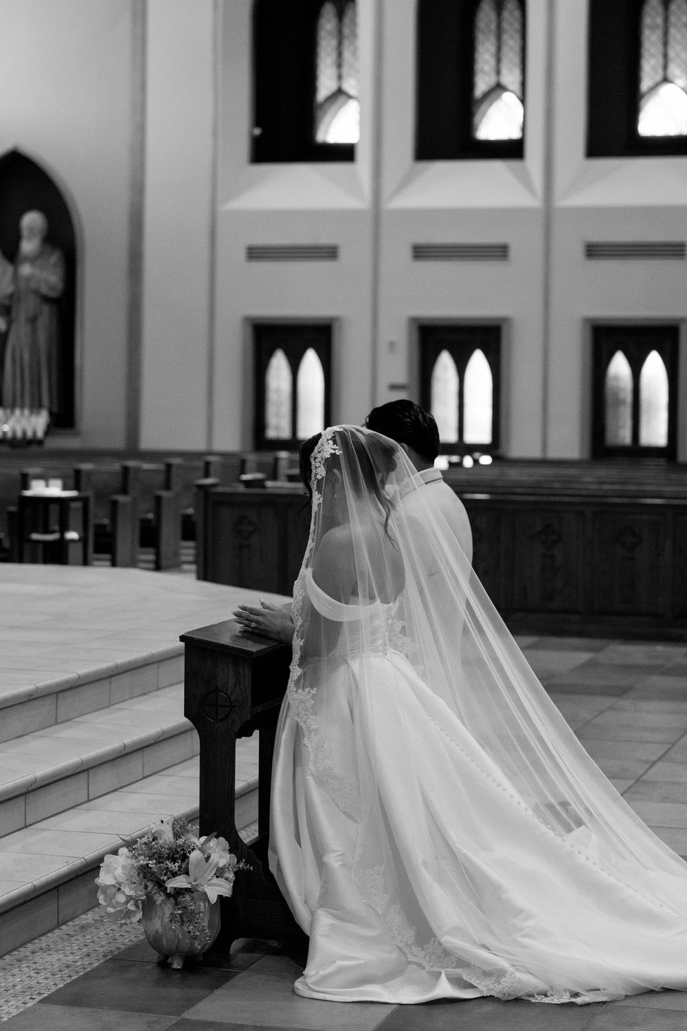 Black and white photo of a bride in an off-the-shoulder gown and veil kneeling with her groom at a church altar.