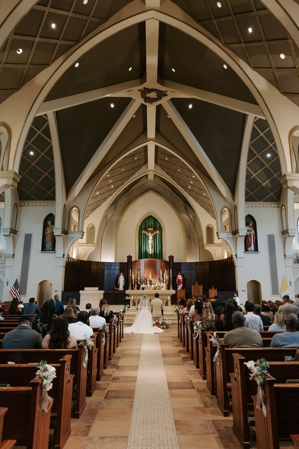 Wide view of a bride and groom at the altar of a grand church as wedding guests fill the pews beneath a vaulted ceiling.