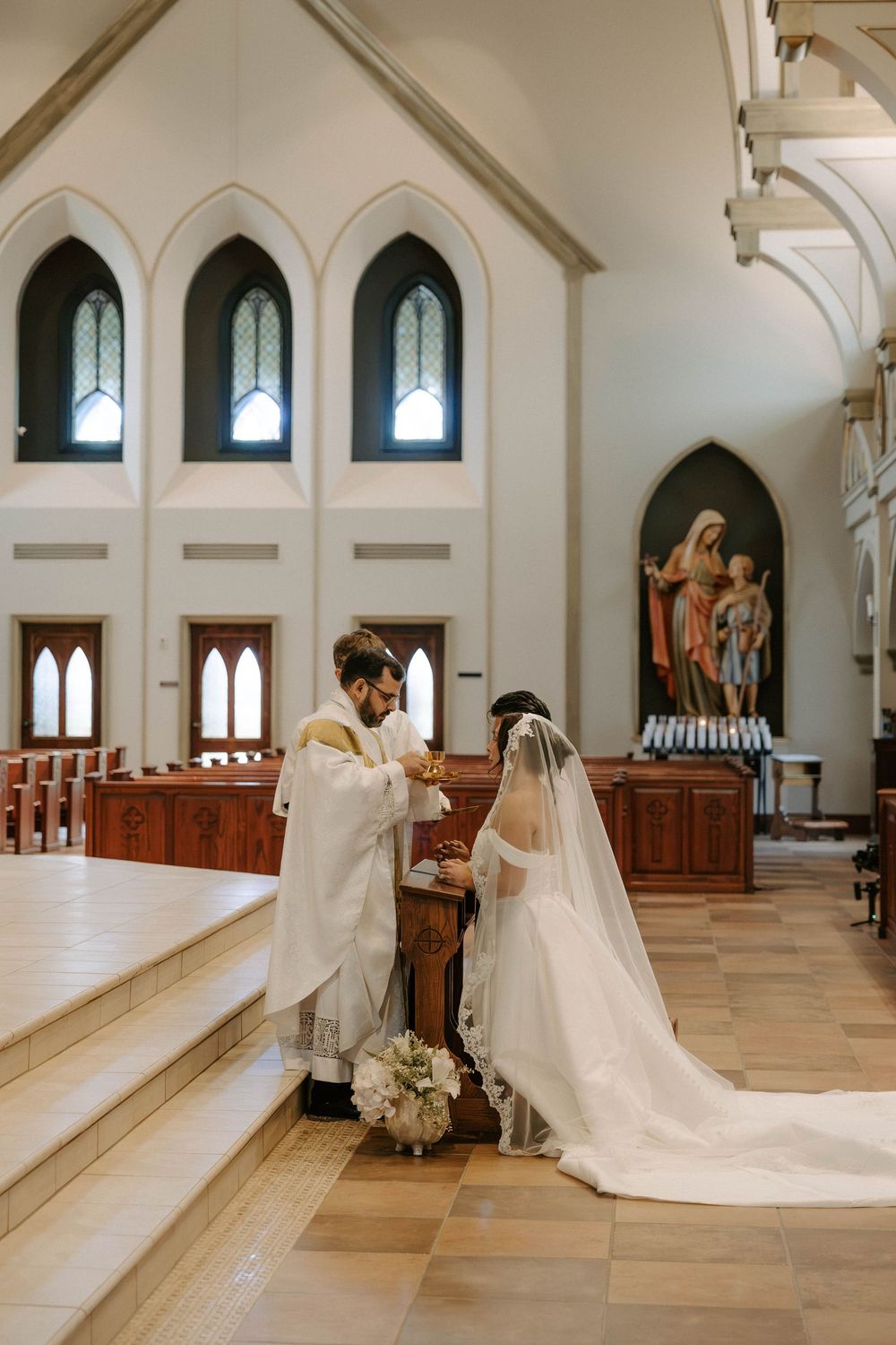 Bride and groom kneel at a prie-dieu while a priest in white vestments blesses them during a Catholic wedding ceremony.