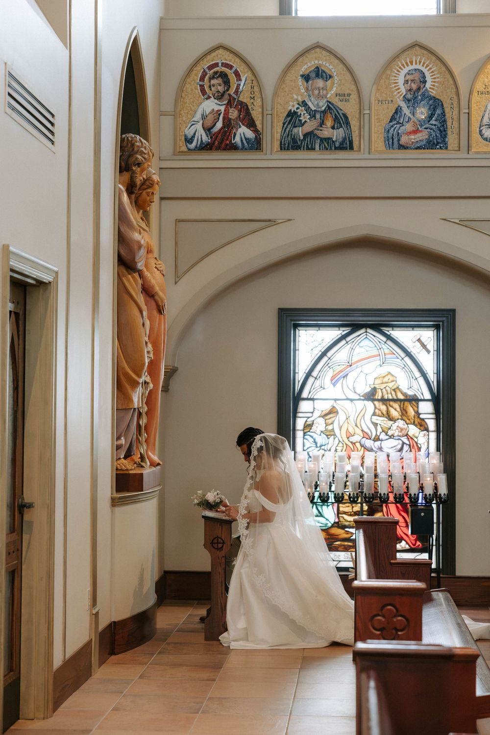 Bride in a white gown and veil kneels in prayer at a church altar rail beneath stained glass and religious icons.
