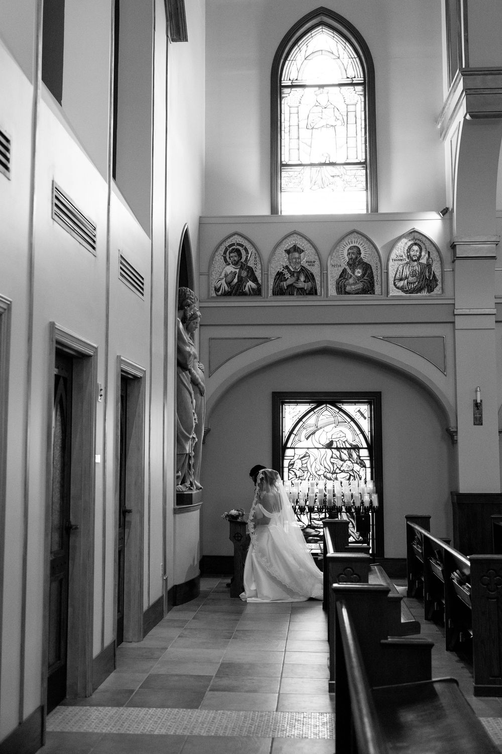Black and white photo of a bride in a long veil walking through a church beneath stained glass windows and saint paintings.