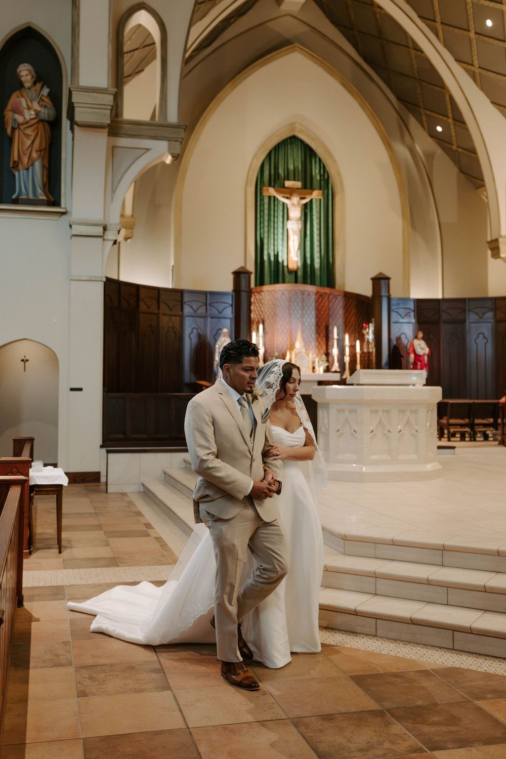 Bride and groom walk hand in hand up the steps of a church sanctuary toward the altar and crucifix.
