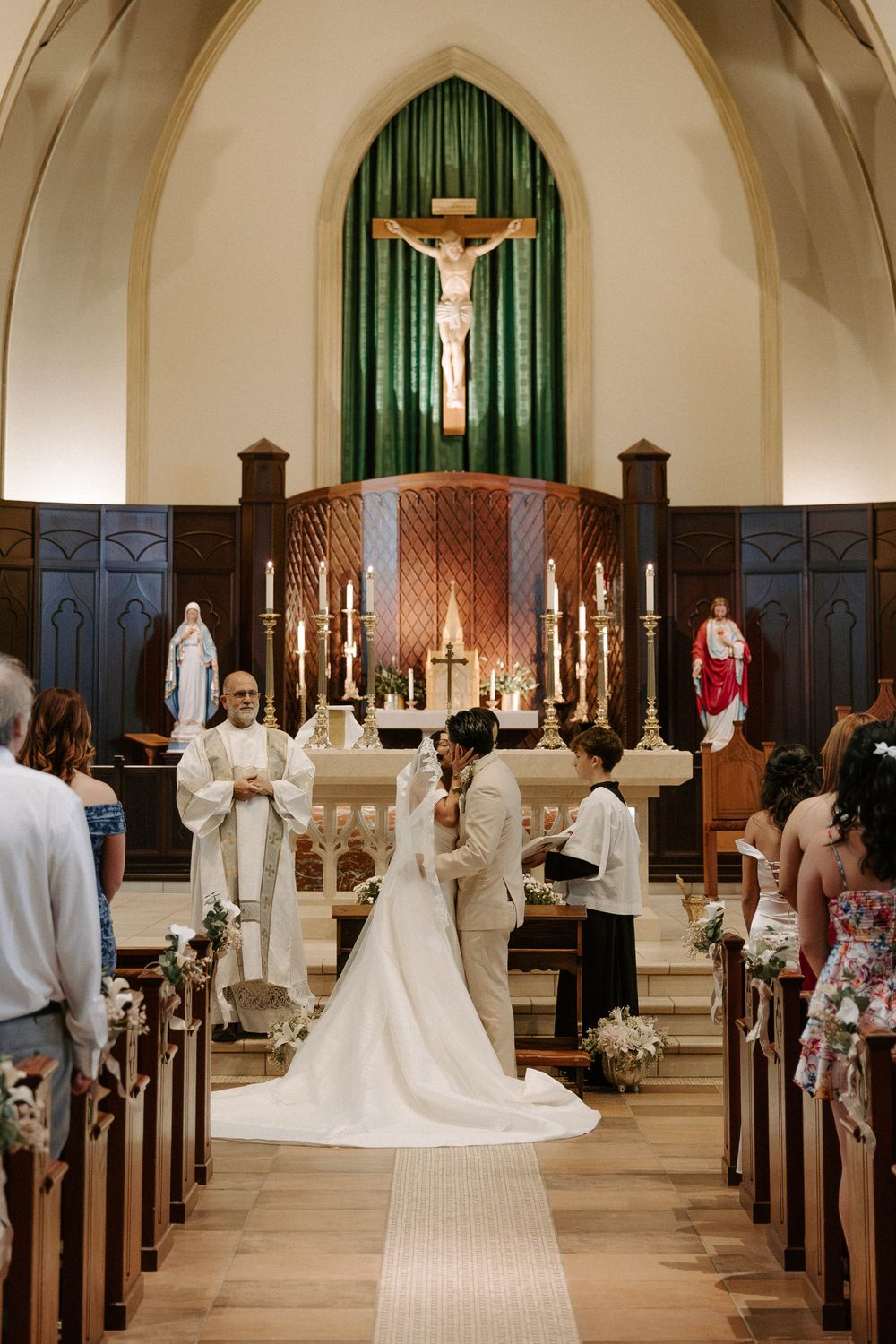 Bride and groom embrace at the altar before a priest and guests during a Catholic wedding ceremony beneath a crucifix.
