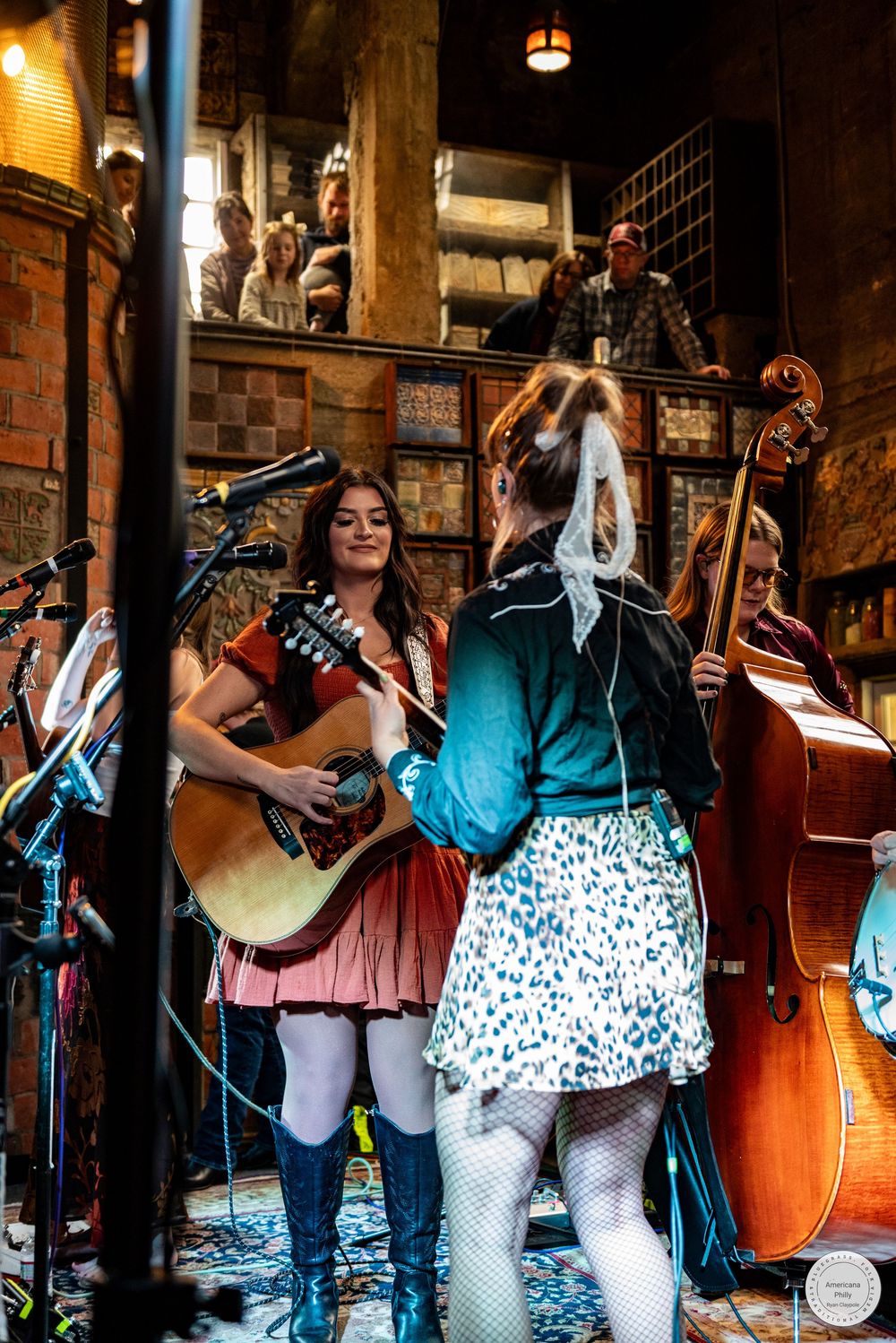 Female bluegrass band performs on stage in a rustic brick venue, with a guitarist, an upright bassist, and an audience watching behind.