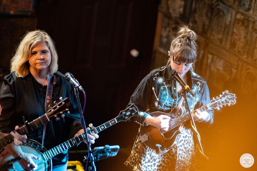 Two musicians perform on stage with stringed instruments, one with short blonde hair and one with a braided updo, singing into microphones.