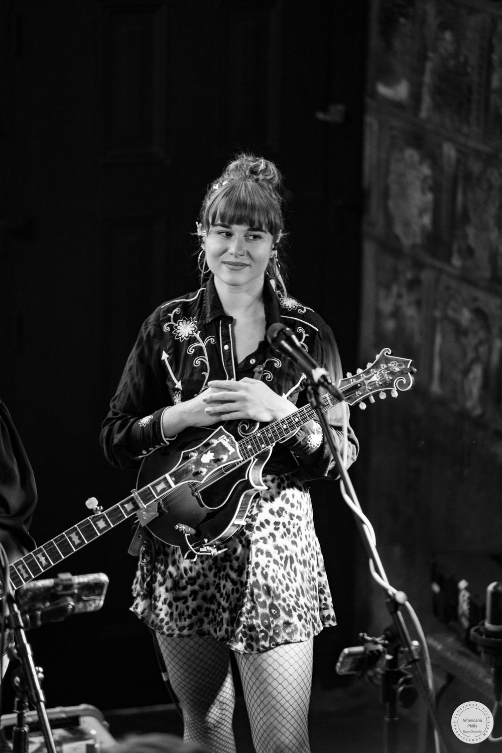 Black and white photo of a smiling mandolin player in an embroidered western shirt and leopard print skirt performing on stage.