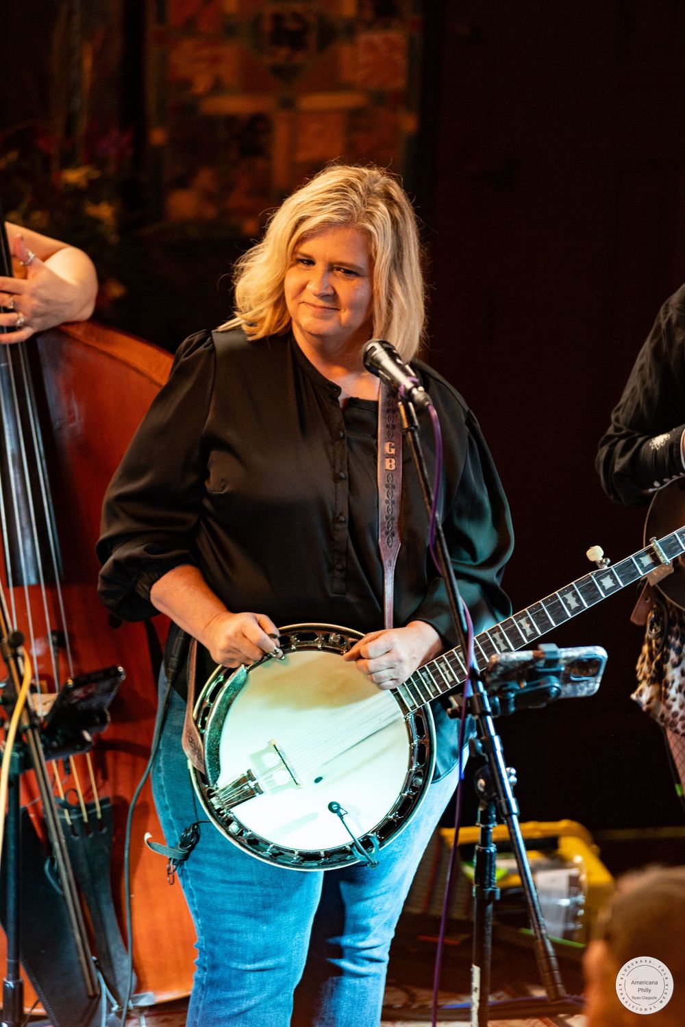 Blonde woman in a black blouse and jeans plays a banjo and sings into a microphone on stage beside an upright bass.