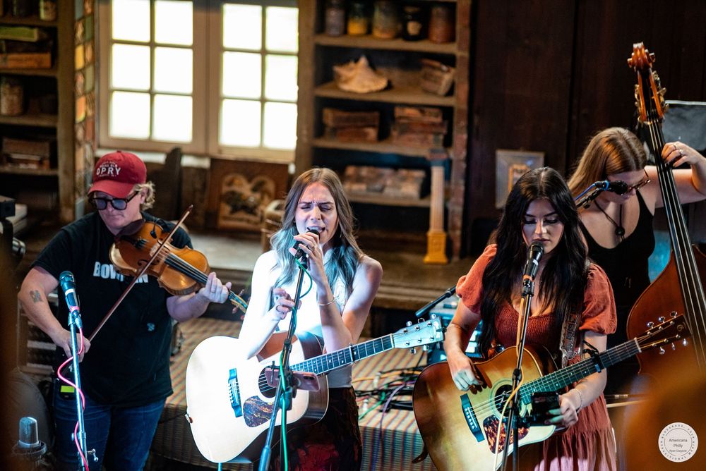 Four musicians perform acoustic folk music on a rustic stage, playing fiddle, guitars and upright bass while singing into microphones.
