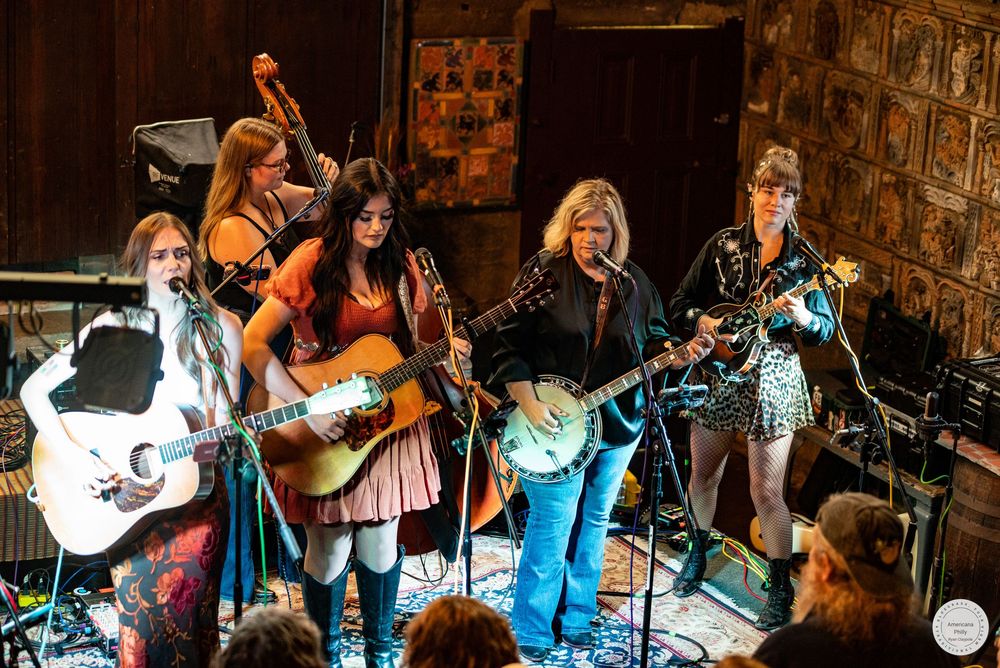 Five women perform on stage in a rustic venue, playing acoustic guitars, banjo and upright bass while an audience watches in the foreground.