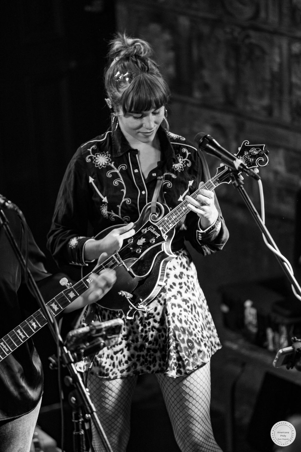 Black and white photo of a woman in a embroidered western shirt and leopard print skirt playing mandolin on stage beside a microphone.