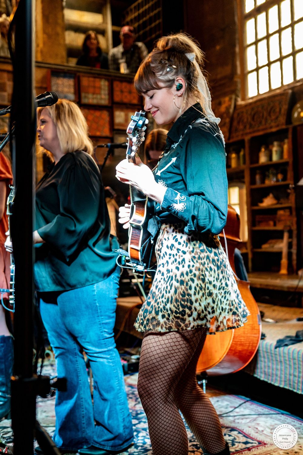 Woman in a leopard print skirt and fishnet tights plays guitar on stage beside a bandmate in a cozy, bookshelf-lined venue.