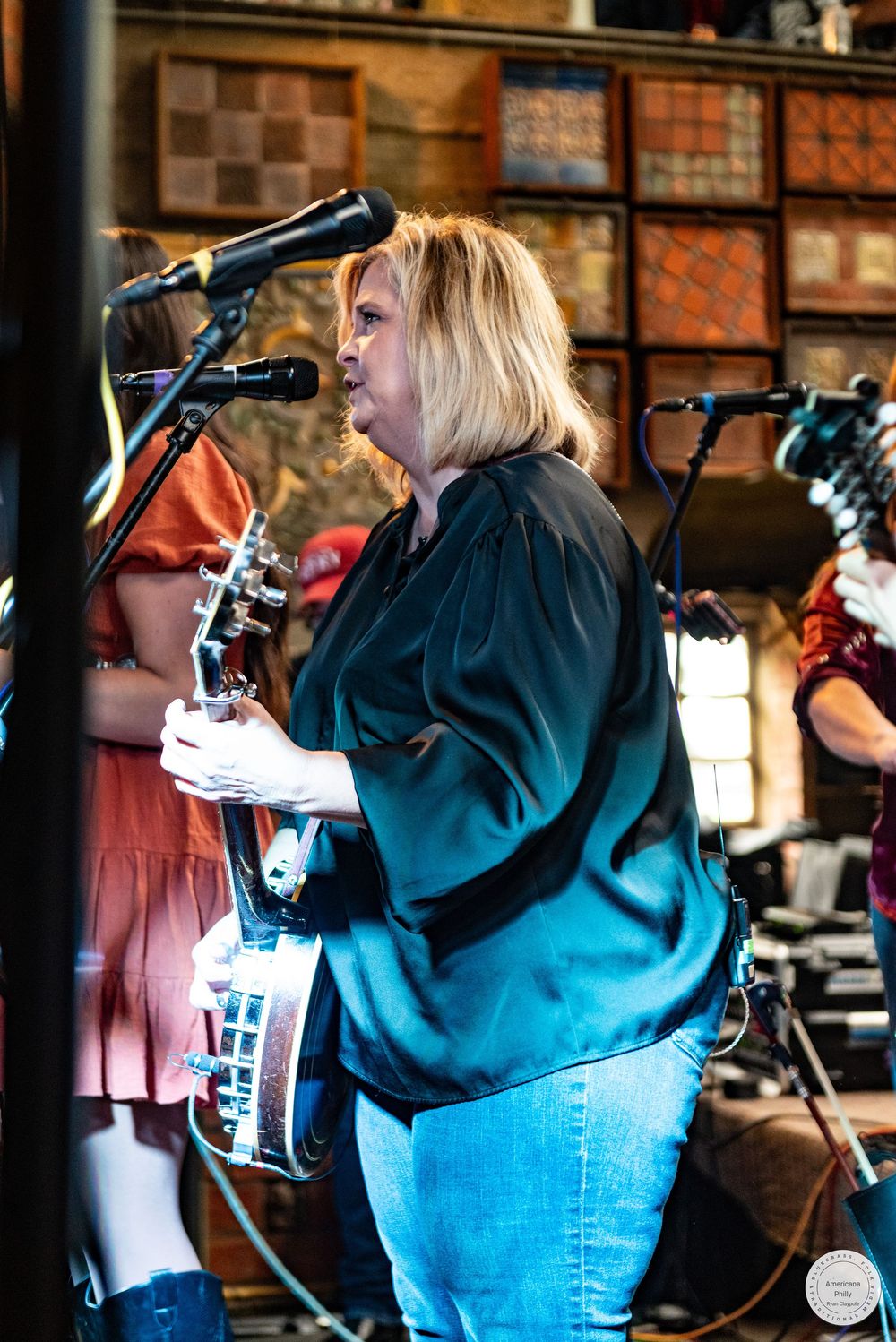 A blonde woman in a teal blouse and jeans plays banjo at a microphone during a live bluegrass performance on stage.