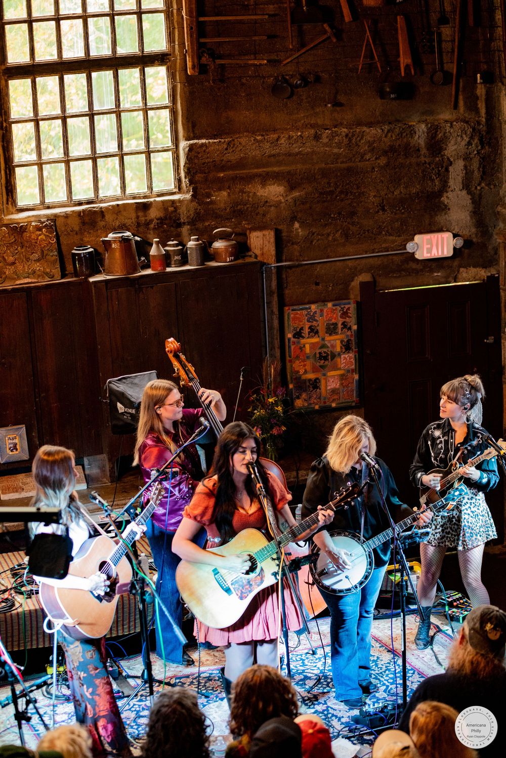 Five musicians play guitars, banjo and fiddle on a rustic stage as an audience watches during an intimate live folk concert.