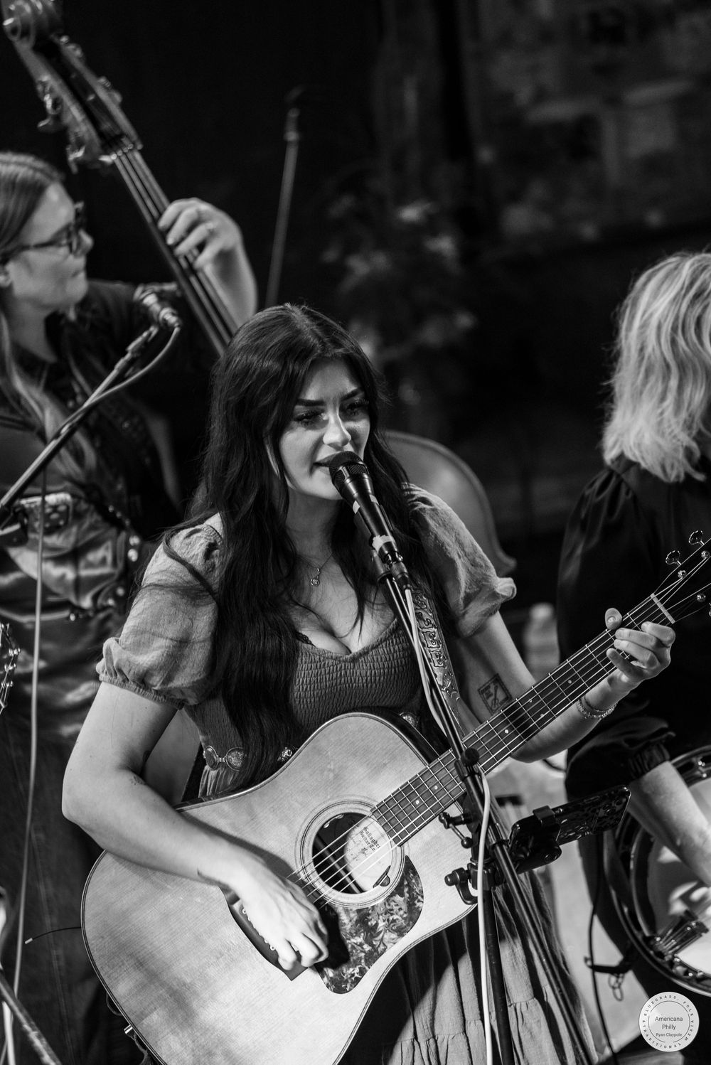 Black and white photo of a long-haired woman singing into a microphone while playing acoustic guitar on stage with bandmates behind her.