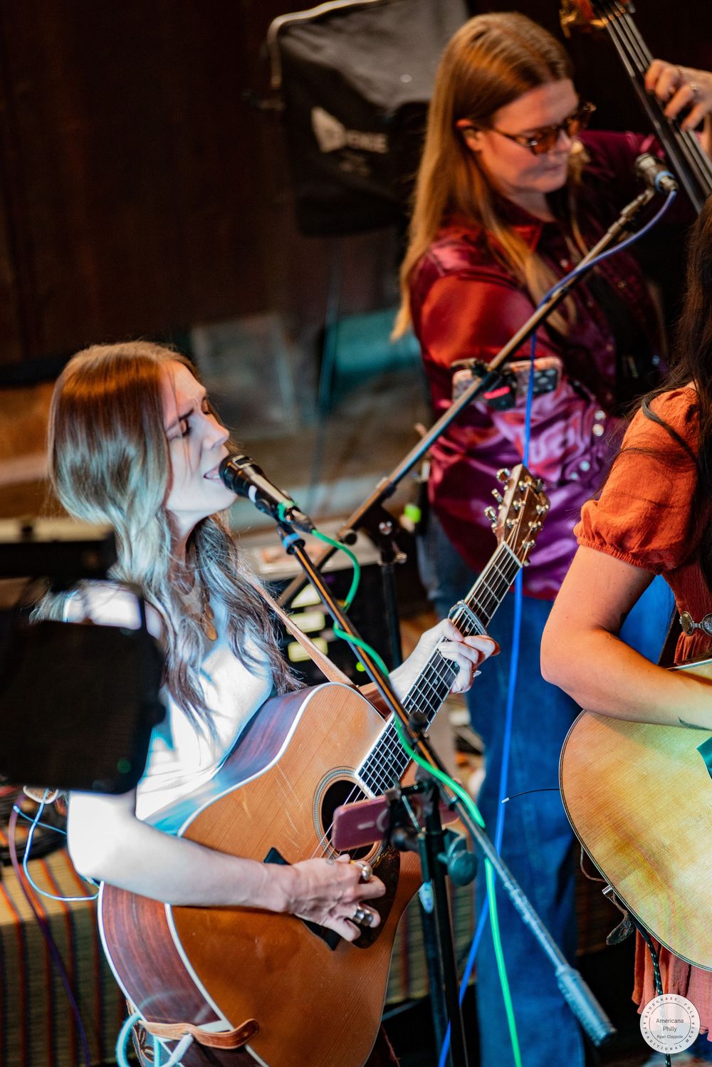 A young woman with long hair sings and plays acoustic guitar at a microphone while bandmates perform behind her at a live concert.
