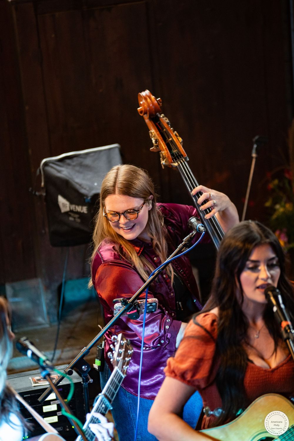 Two women perform on stage, one smiling while playing an upright bass behind a microphone and another singing with an acoustic guitar.