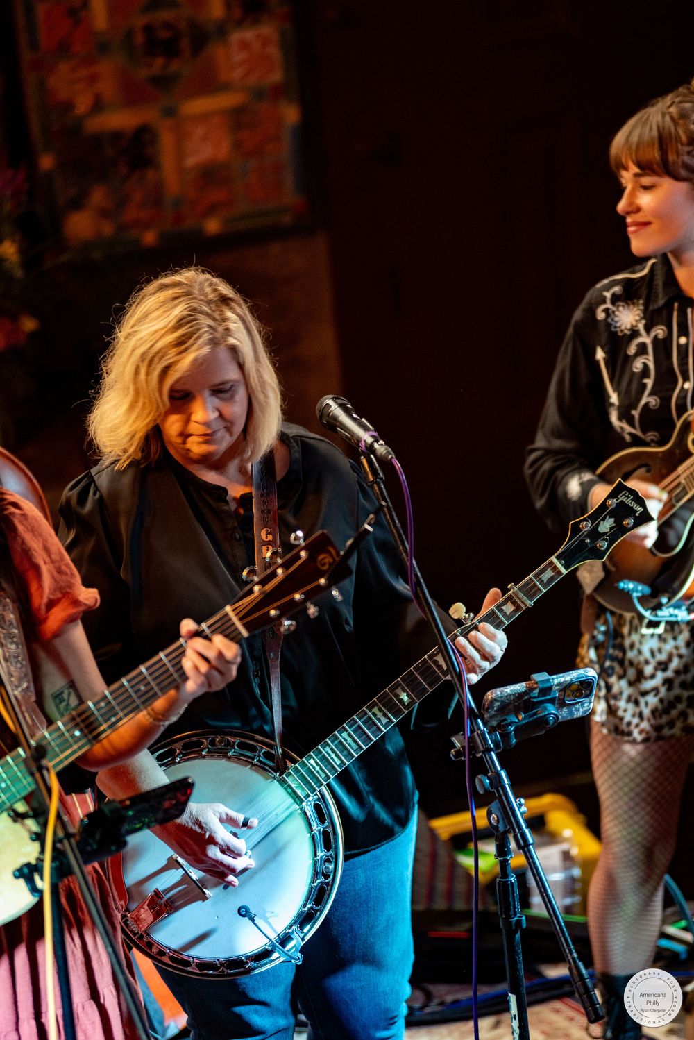 Blonde musician plays a banjo at a microphone on stage beside a smiling bandmate in a western-style embroidered shirt holding a guitar.