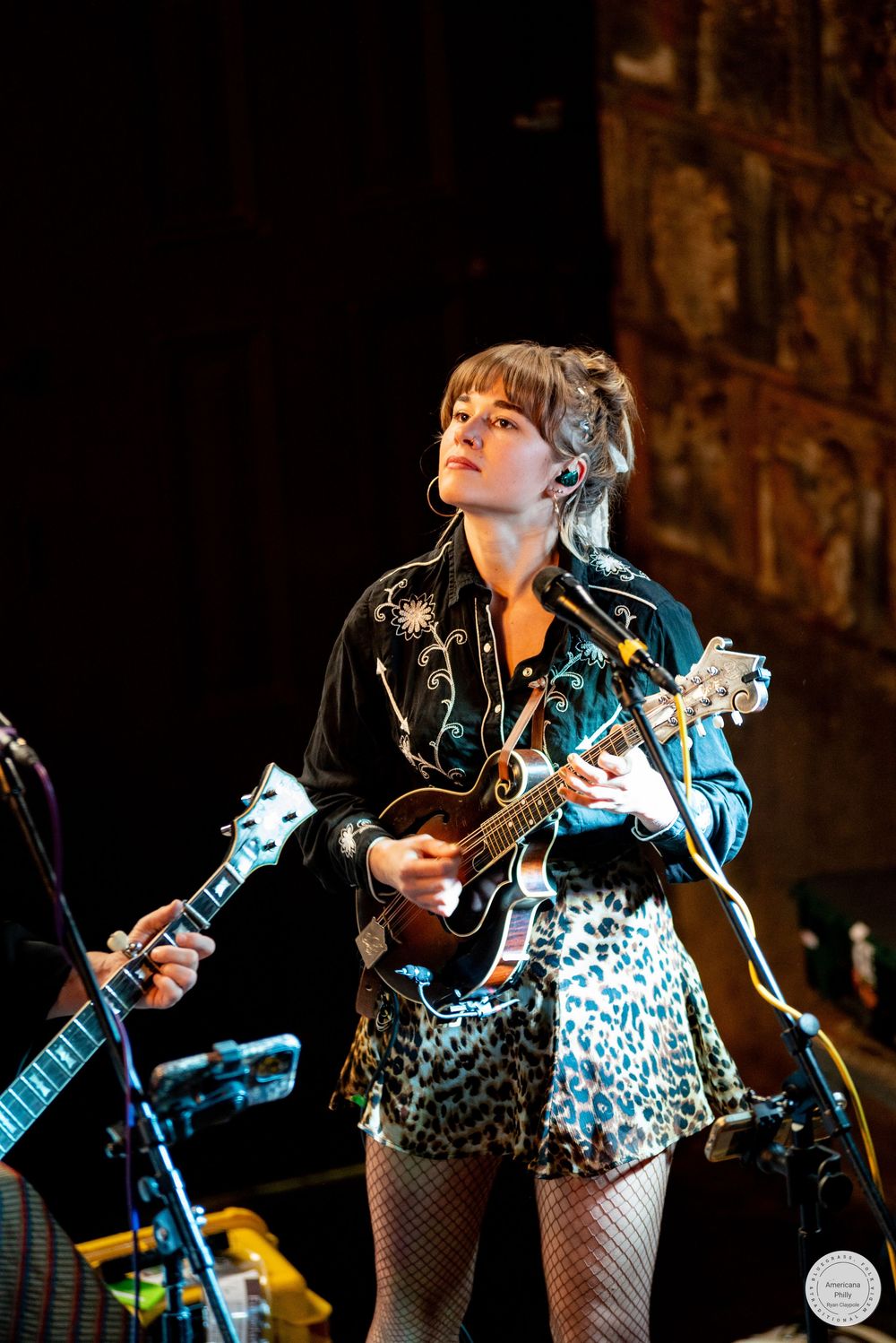 A woman in a western-style embroidered shirt and leopard print skirt plays mandolin on stage under dramatic concert lighting.