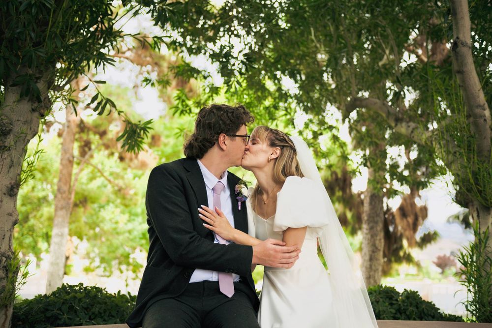 A bride in a white gown and veil shares a kiss with her groom in a dark suit while seated outdoors beneath leafy green trees.