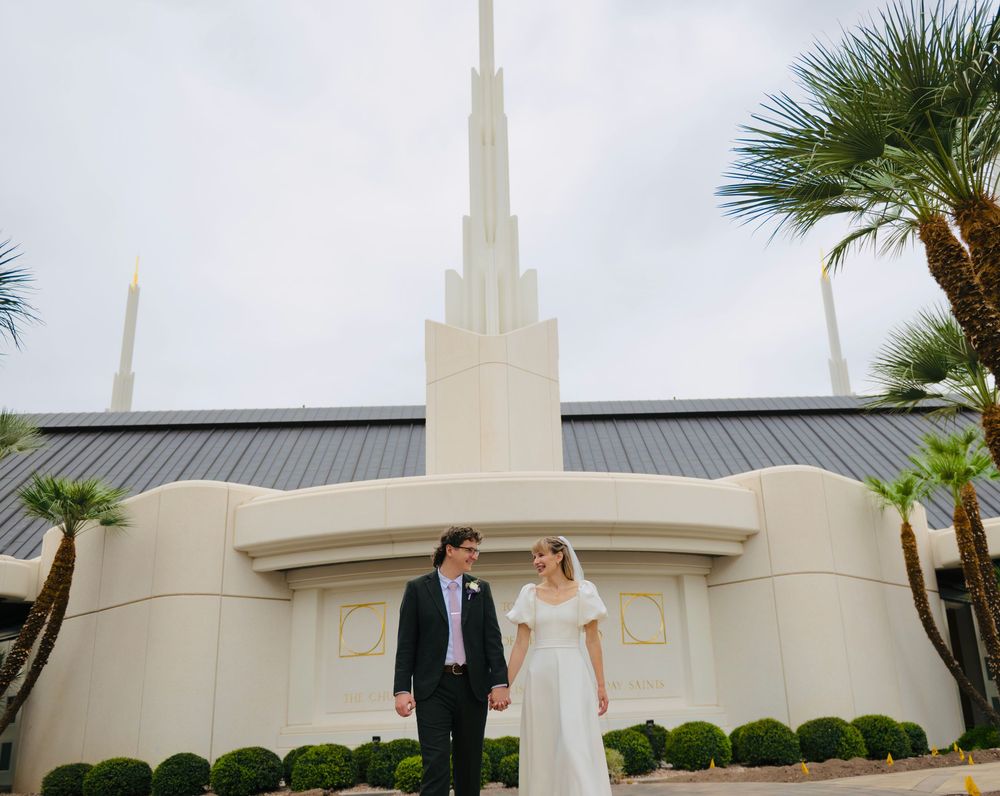 A bride and groom hold hands outside a white temple with a tall spire, surrounded by palm trees and trimmed hedges under a cloudy sky.