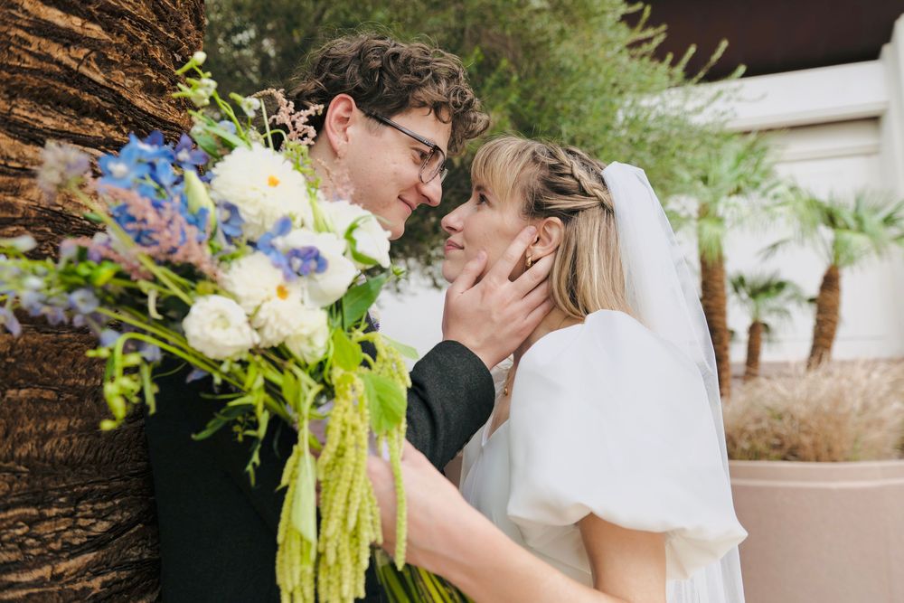 A smiling groom in glasses gently touches his bride's face beside a colorful bouquet of white and blue flowers.