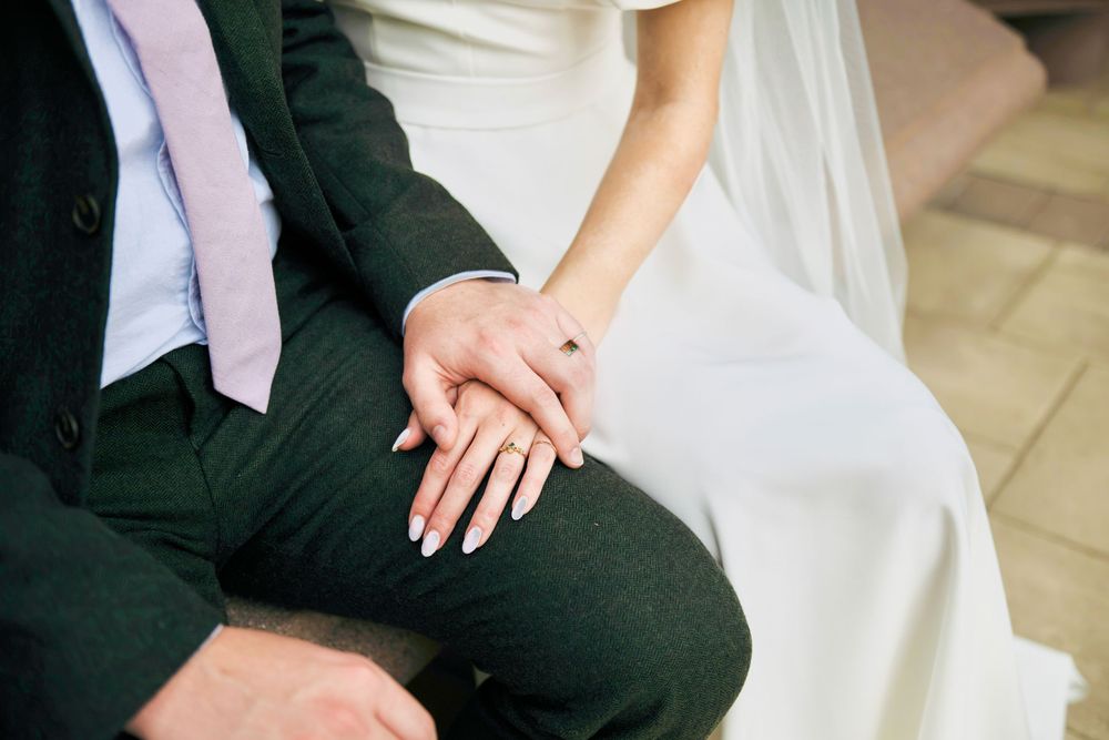 Bride in a white gown and groom in a dark green suit sit together, her hand with a ring resting gently on his as they hold hands.