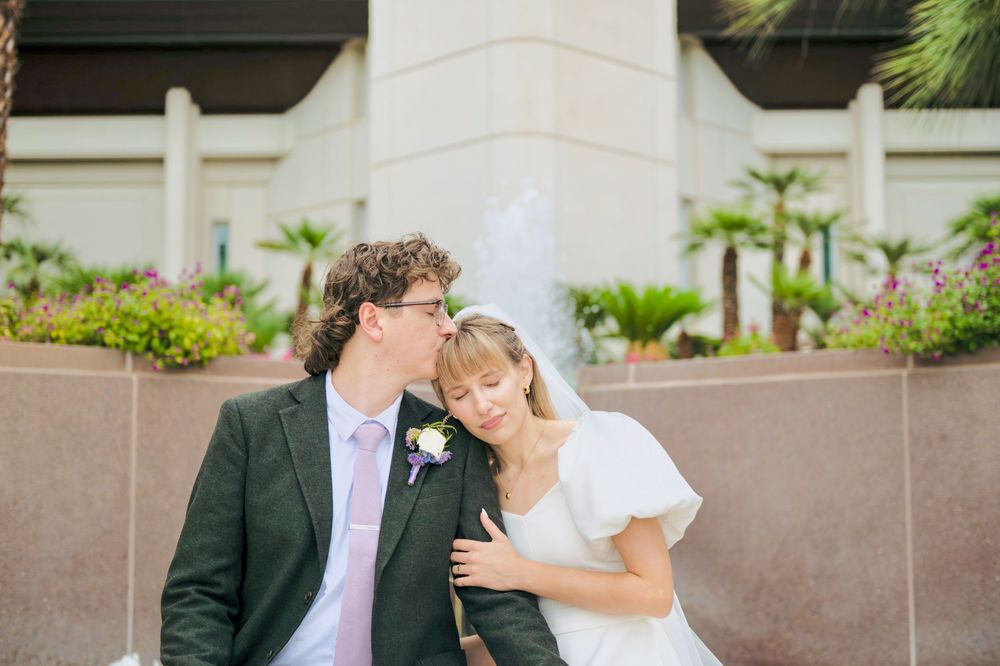 Smiling bride in a white dress rests her head on the shoulder of a groom in a gray suit and pink tie near palm trees and a fountain.
