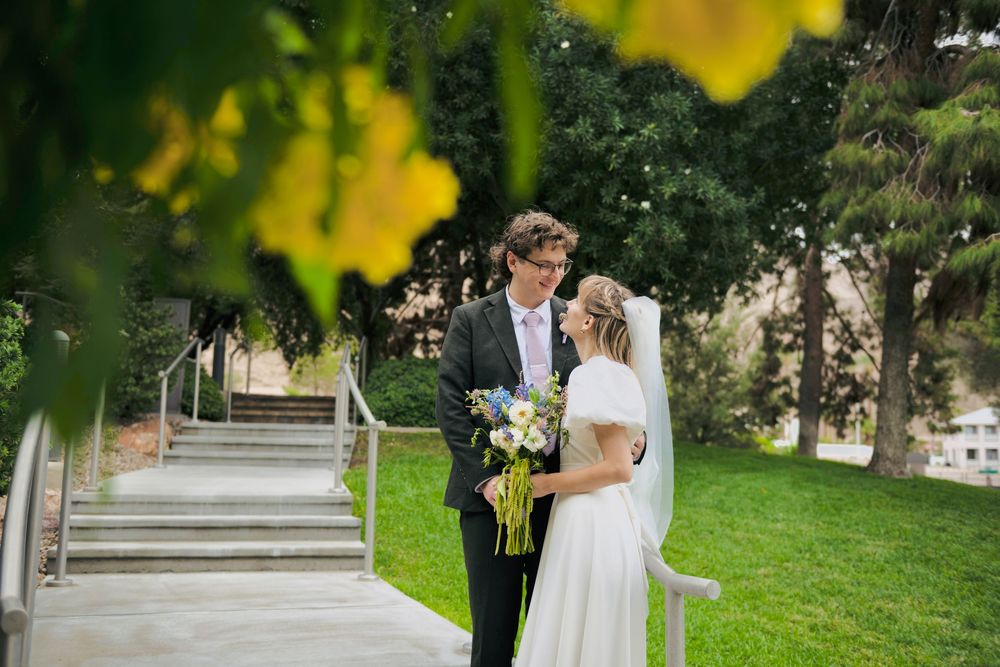 Bride in a white gown holding a bouquet embraces her groom in a dark suit on a sunny lawn beside a stone staircase surrounded by trees.
