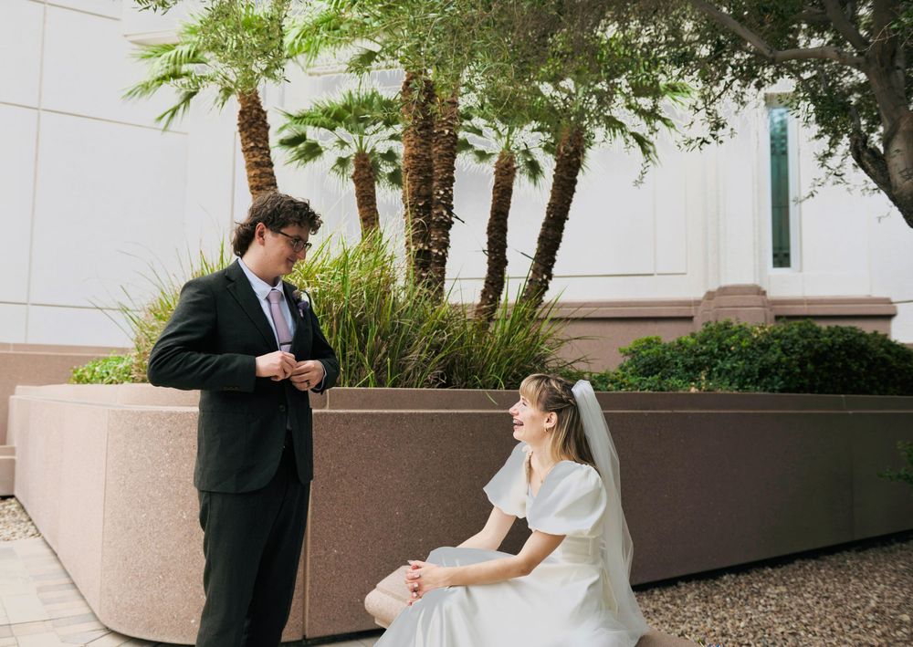 Bride in a white gown and veil sits on a stone planter while the groom in a dark suit stands beside her near palm trees.