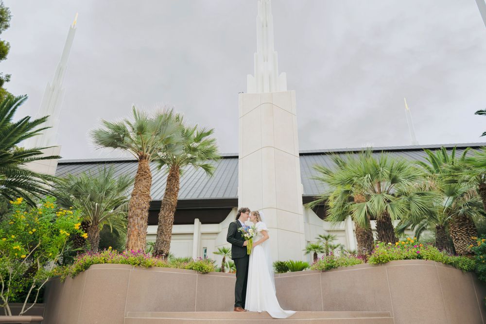 Bride in a white gown and groom in a dark suit embrace on temple steps surrounded by palm trees and a tall white spire under a cloudy sky.