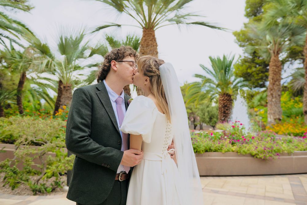 Bride in a white dress and veil kisses a groom in a dark suit and purple tie in a sunny garden with palm trees and flowers.