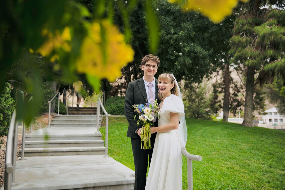 Smiling bride in a white dress and groom in a dark suit hold a bouquet on a green lawn beside stone steps, framed by yellow flowers.