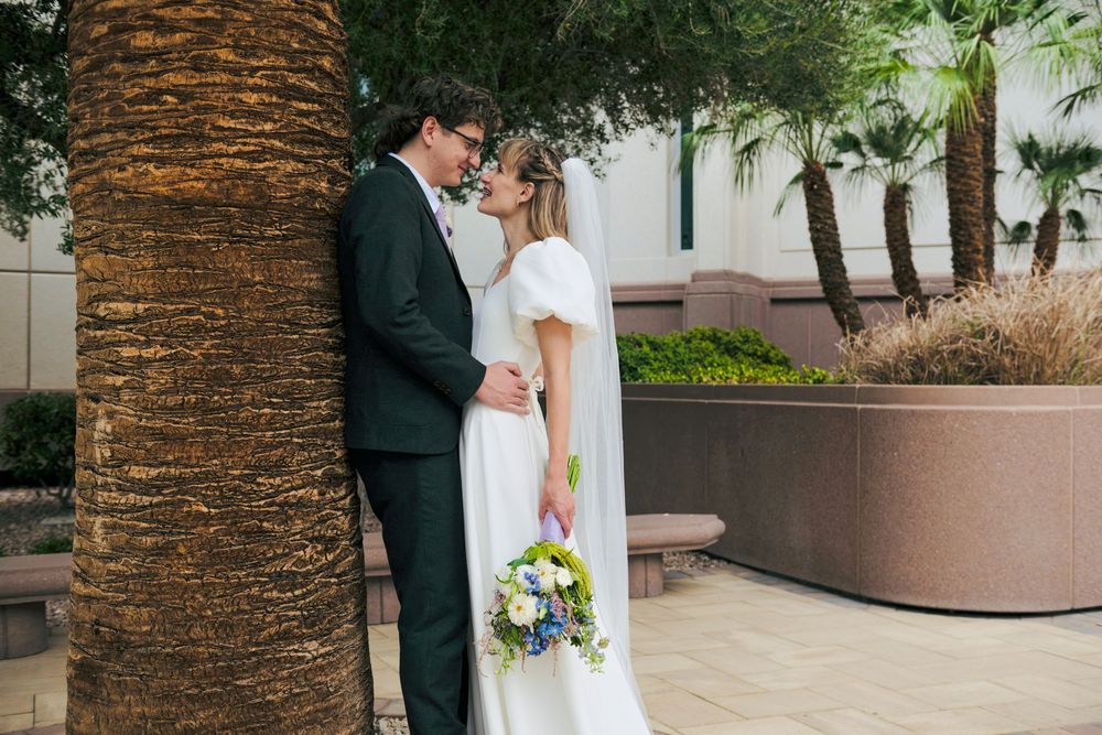 Bride in a white dress holding a colorful bouquet embraces her groom in a dark suit beside a palm tree in a sunny courtyard.