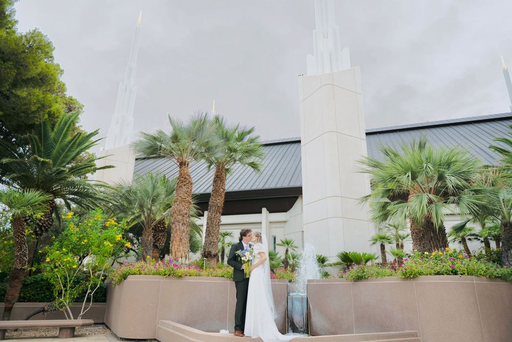 A bride in a white gown and a groom in a dark suit embrace beside palm trees outside a temple with tall white spires.