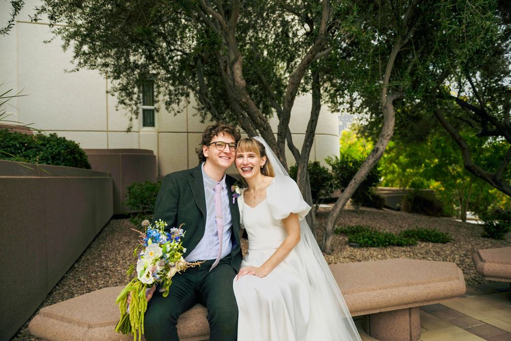 A smiling bride in a white gown and veil sits beside a groom in a dark suit on a stone bench, holding a colorful bouquet.