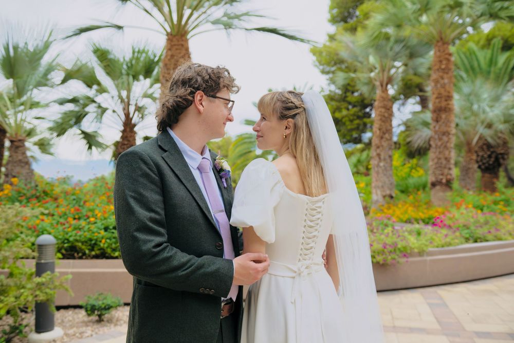 A bride in a white lace-back wedding dress and veil smiles at her groom in a dark suit among palm trees in a sunny garden.