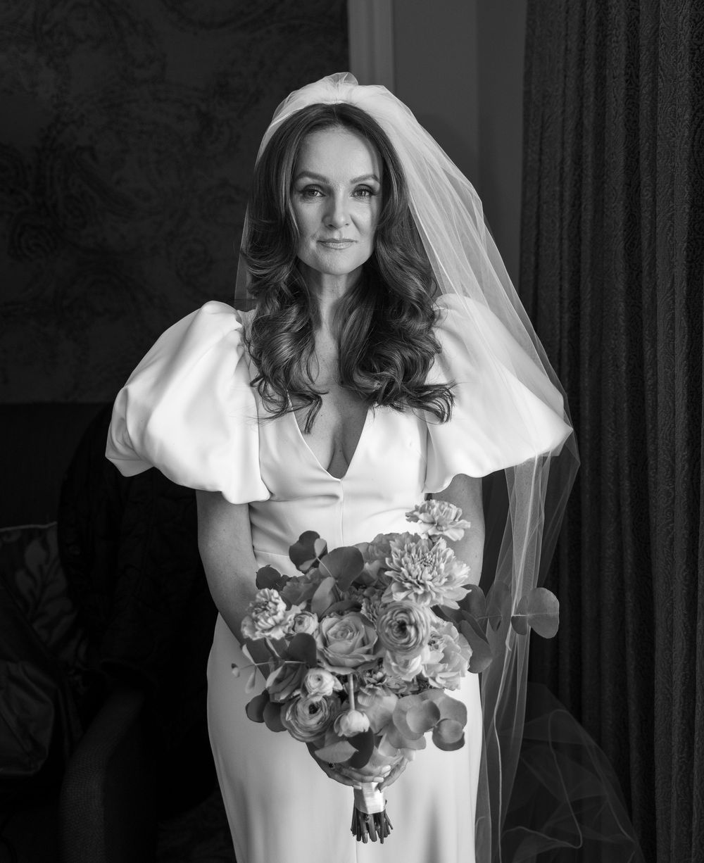 Elegant bride in white gown with puff sleeves and veil holds floral bouquet in black and white portrait photo.