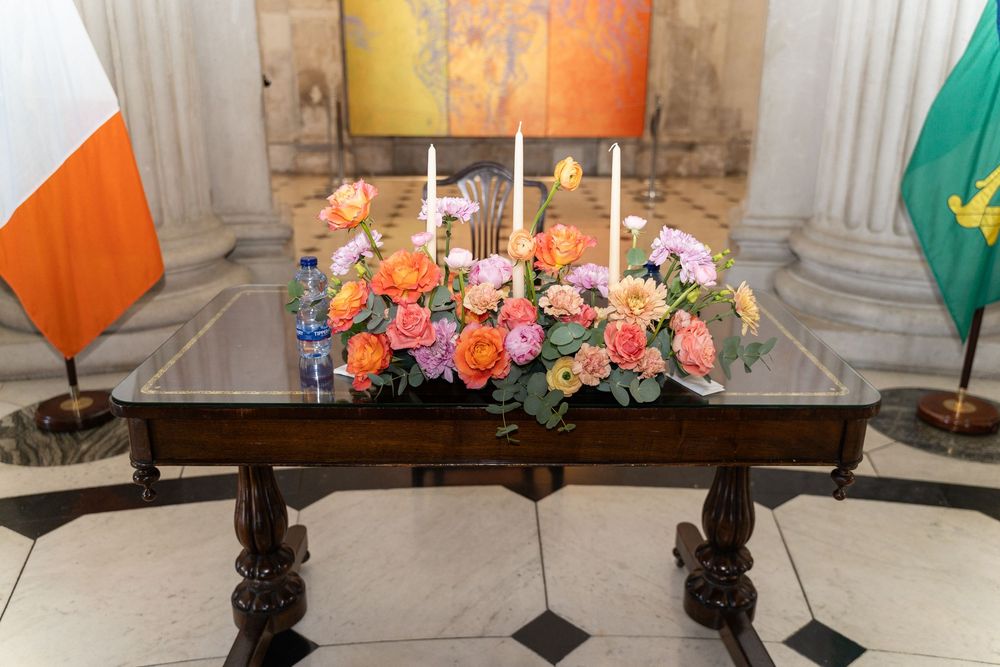 Elegant floral arrangement with orange and pink roses, white candles on a wooden table flanked by Irish and green flags.