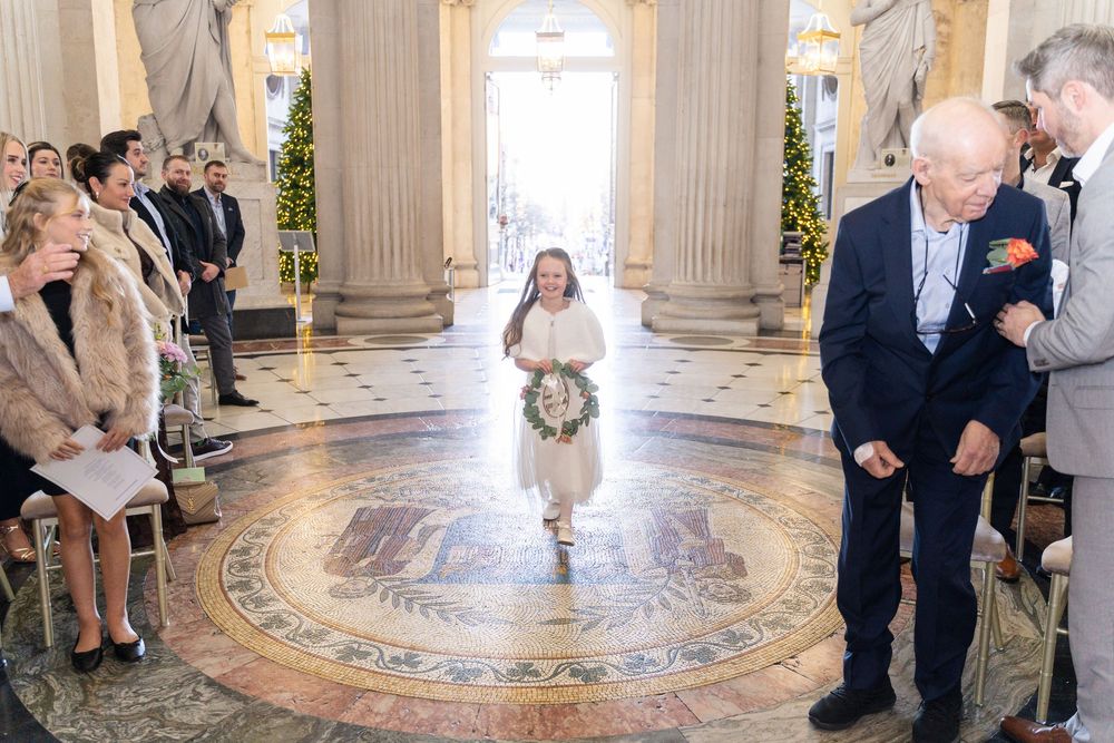 Young flower girl walks down ornate wedding aisle in white dress, carrying wreath, as guests watch in elegant marble hall.
