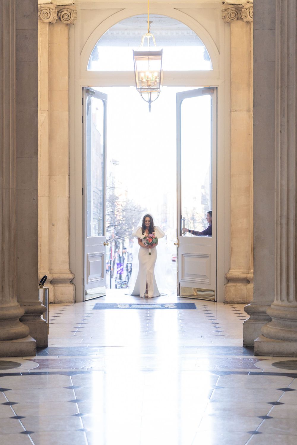 Bride in white fitted gown walks through grand arched doorway of elegant venue holding bouquet.
