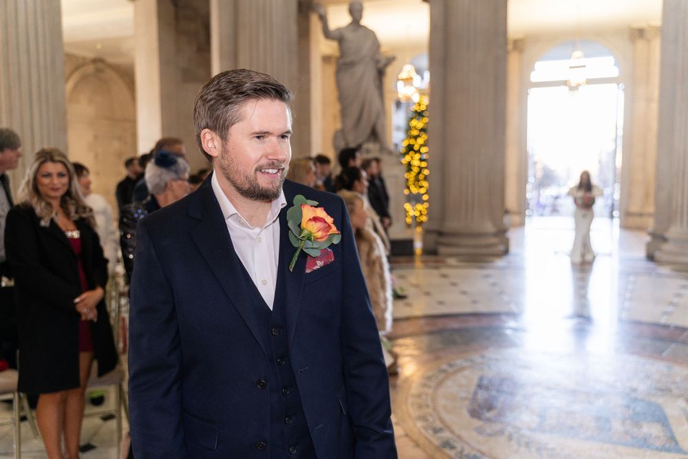 Groom in navy suit with floral boutonniere smiling at wedding ceremony in elegant columned hall.