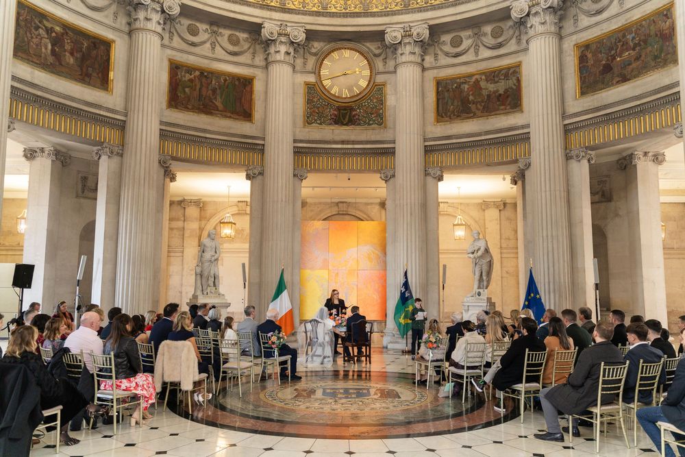 Formal ceremony in a grand neoclassical rotunda with columns, murals, Irish flag, and seated audience on gold chairs.