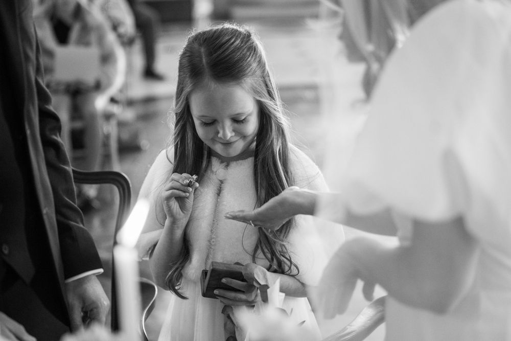 Young flower girl in white dress smiles at phone during wedding ceremony, black and white photo.