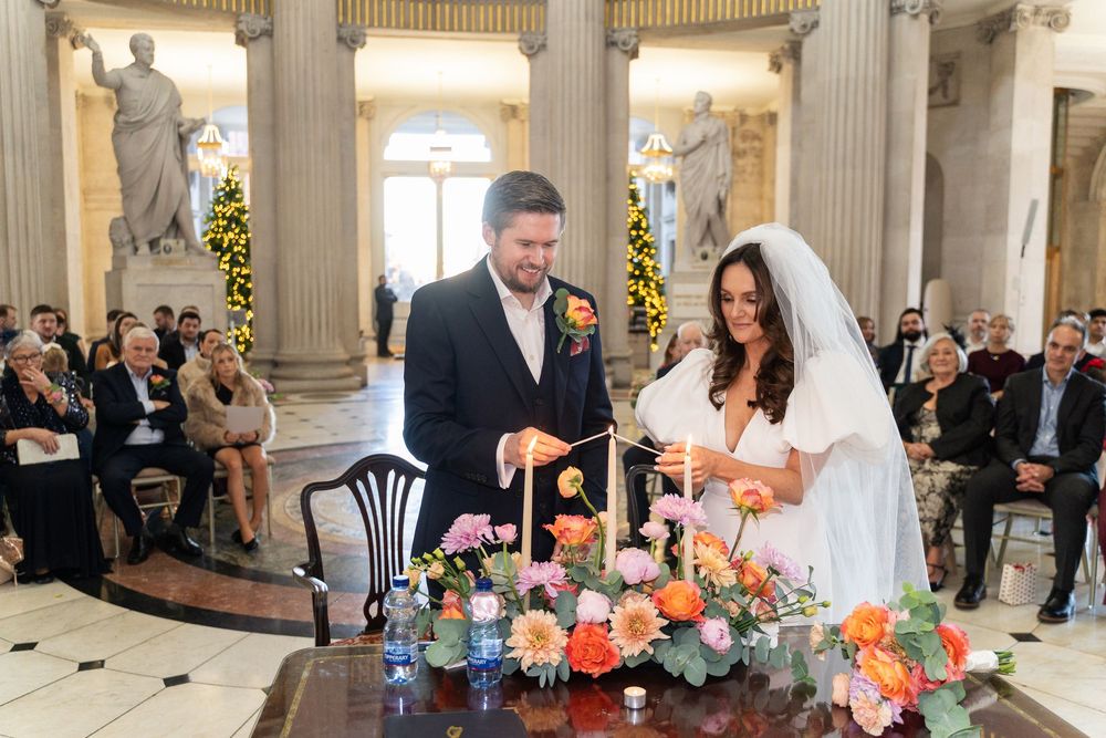 Bride and groom lighting a unity candle at wedding ceremony altar with colorful floral arrangement.