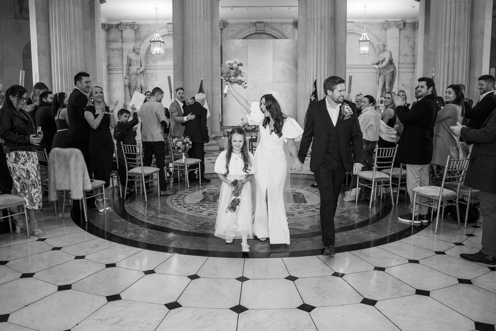 Black and white photo of a bride and groom walking down the aisle with a flower girl at an elegant wedding ceremony.