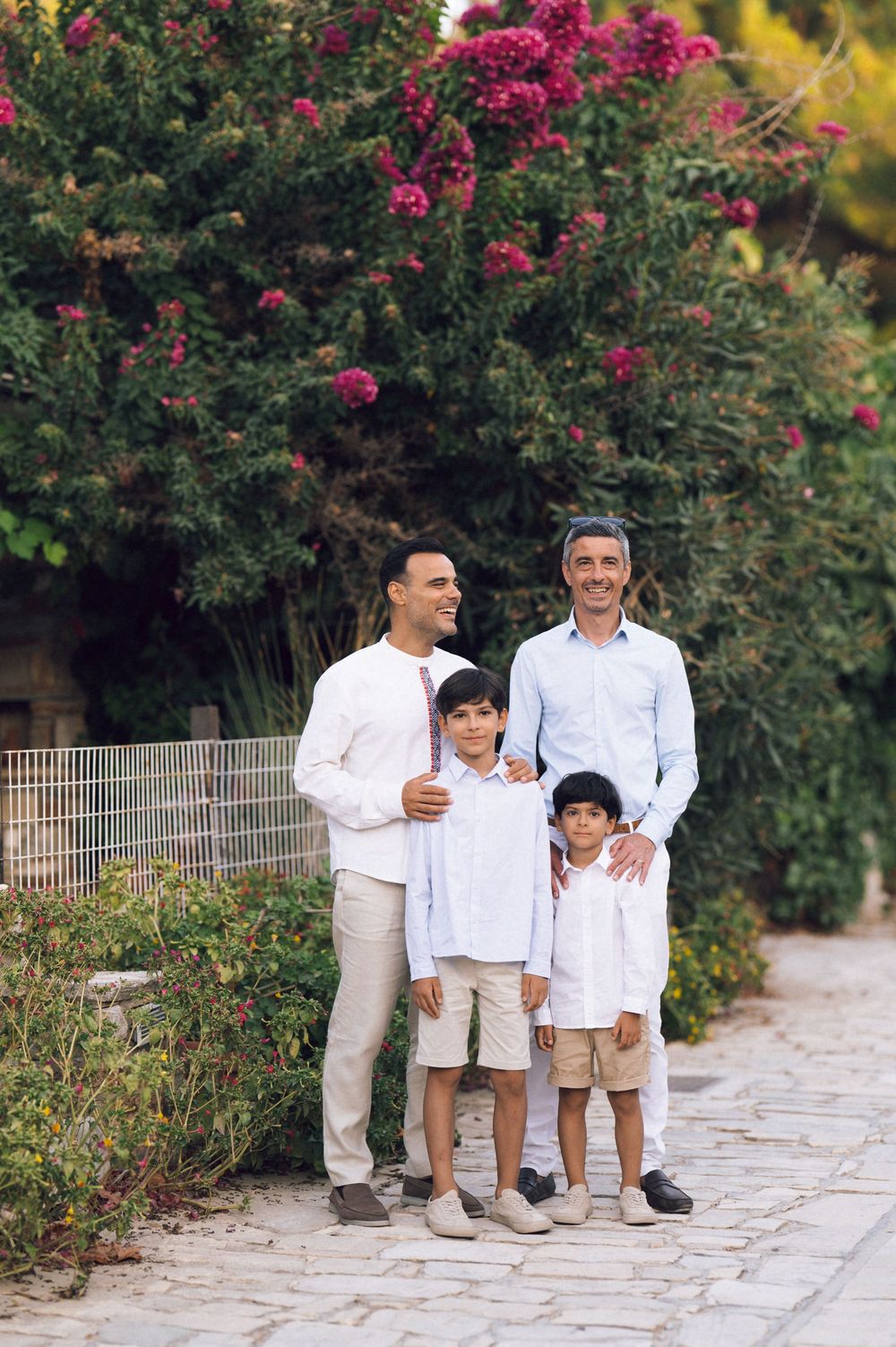 Two men and two boys pose together on a stone path surrounded by lush green shrubs and pink flowering trees.