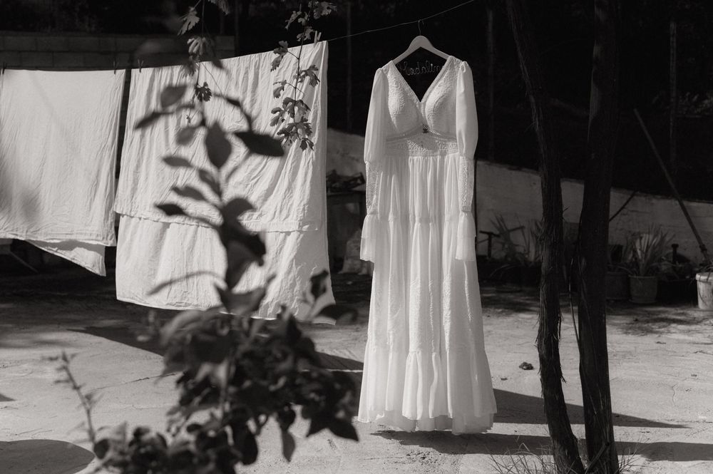 Black and white photo of an elegant white wedding dress hanging on a clothesline outdoors among trees.