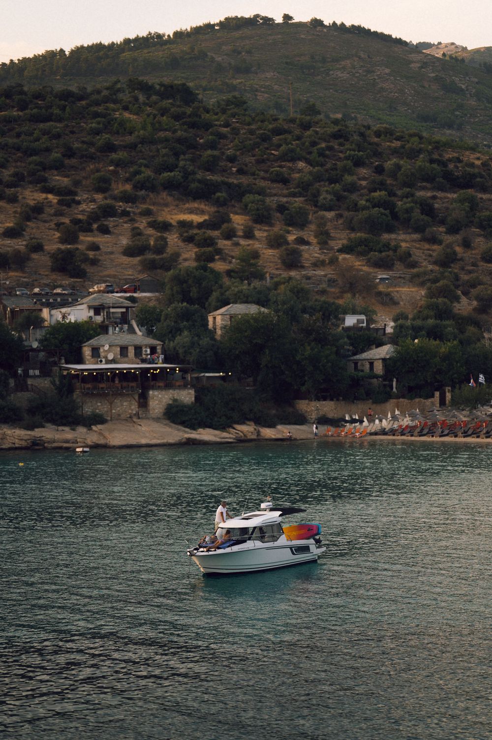 A small white motorboat cruises on turquoise waters near a hillside village with lush green trees at dusk.
