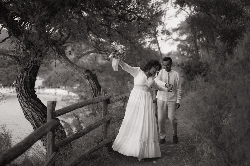 Bride in white gown dancing with groom on a scenic tree-lined path with wooden fence in black and white.