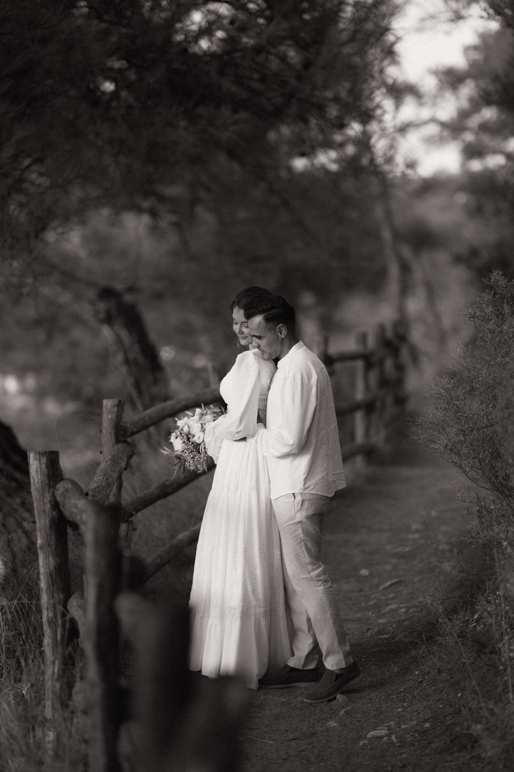 Black and white photo of a couple in white attire embracing on a rustic path surrounded by trees.